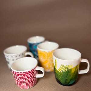 May include: A collection of five ceramic mugs featuring distinct patterns and colors. One mug displays a red scalloped design, another a green and yellow abstract brushstroke print, a third a yellow floral motif, and two others with blue and black patterns.