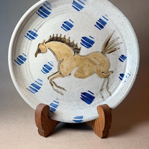 May include: A round ceramic plate with a speckled off-white glaze featuring a hand-painted tan horse in motion at the center. The plate is decorated with scattered blue brushstroke patterns around the horse and rests on a wooden stand.