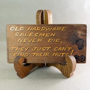 May include: A rectangular wooden sign resting on a wooden stand. The sign features the engraved text: OLD HARDWARE SALESMEN NEVER DIE, THEY JUST CAN'T FIND THEIR NUTS! Two metal nuts are attached to the bottom right corner of the wooden board.
