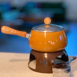 May include: An orange fondue pot with a wooden handle and lid, sitting on a brown base. A small, silver container is next to the pot. The pot has a glass lid and a wooden knob.