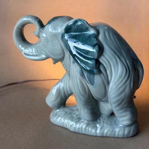 May include: A ceramic elephant figurine in shades of gray and teal. The elephant is posed walking, with its trunk raised and ears spread. The figurine has a glossy finish and is mounted on a textured base.