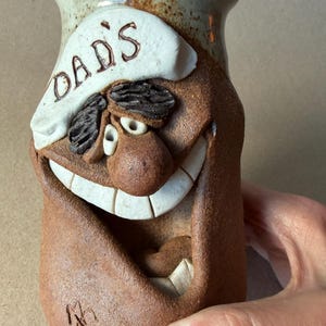 May include: A brown ceramic mug with a smiling face design. The mug features a white chef's hat with the word "DAD'S" and a large, open mouth with white teeth. The mug is held by a hand.