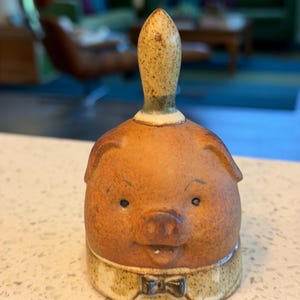 May include: A ceramic pig-shaped bell with a brown body, a beige collar, and a dark bow tie. The bell has a long, speckled handle and small, dark eyes. The bell is a decorative item.
