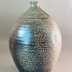 May include: A ceramic vase with a rounded, bulbous body and a narrow neck. The surface features a textured, speckled glaze that transitions from a dark, deep brown at the base to a lighter, cream-colored finish toward the top, with visible horizontal ridges.