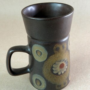 May include: A tall, dark brown ceramic mug with a flared rim and a single handle. The exterior features a repeating pattern of light green circles and a central stylized floral motif in shades of tan, brown, and a small dark red center point.