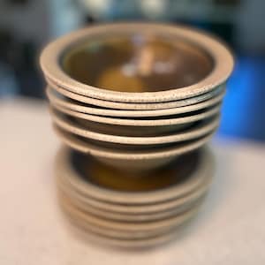 May include: A stack of beige ceramic bowls with a wide rim and a slightly textured surface. The bowls have a light brown interior and are stacked on top of each other, with a similar stack below.