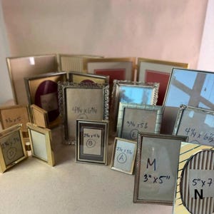 May include: A collection of various metal picture frames in different sizes and finishes, including gold, silver, and bronze tones. Several frames feature handwritten labels with dimensions in inches, such as 3x5, 4.25x6.25, 2.25x4.25, 2.25x3.25, and 5x7.