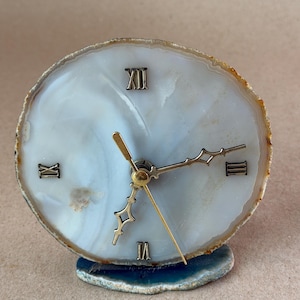 May include: A circular clock face crafted from a translucent white agate slice with natural banding. It features metallic Roman numerals at the twelve, three, six, and nine o'clock positions, with gold-toned clock hands mounted in the center.