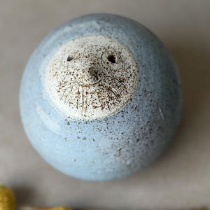 May include: A spherical ceramic object with a light blue speckled glaze. The top features a circular, off-white inset area with a textured, dark brown speckled finish, depicting a simple face with two small eye holes and etched whiskers.