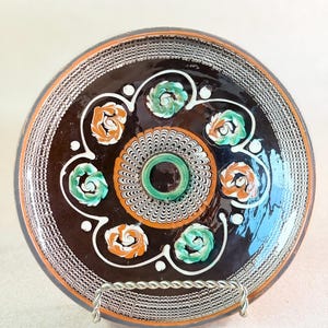 May include: A round ceramic plate with a dark brown glazed surface featuring a central circular pattern of white and orange swirls. The plate is decorated with alternating orange and green floral motifs and a white lattice border pattern.