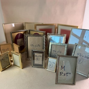 May include: A collection of various rectangular metal picture frames in gold and silver tones, featuring different border designs and sizes. Several frames have handwritten labels indicating dimensions such as 3x5 inches, 4.25x6.5 inches, and 2.25x3.25 inches, along with identifying letters.