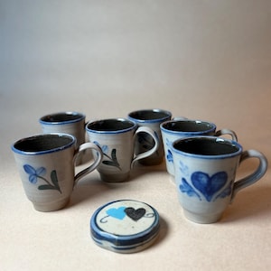 May include: A set of five light gray ceramic mugs with blue painted floral and heart motifs, featuring dark blue rims and handles. A small, round, flat ceramic lid with a painted light blue heart and a black heart sits in the foreground.