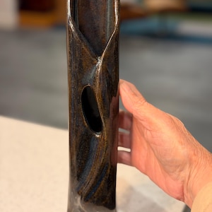 May include: A tall, handcrafted ceramic vase with a dark brown and bronze glaze. The vase has a unique, organic shape with a central opening and textured surface. The vase is approximately 12 inches tall.