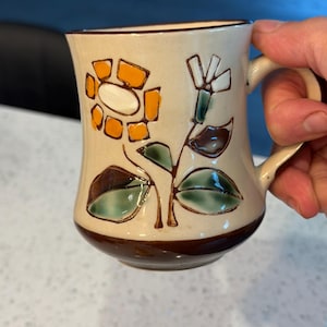 May include: Cream-colored ceramic mug with a brown base and rim, featuring a floral design. The flowers are orange and white, with green and brown leaves. The mug has a curved handle.