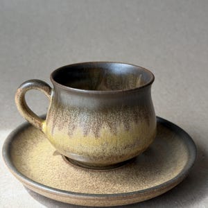 May include: A ceramic coffee cup and saucer set. The cup has a rounded shape with a handle and is placed on a matching saucer. The set features a gradient of colors, transitioning from dark brown to yellow and tan.