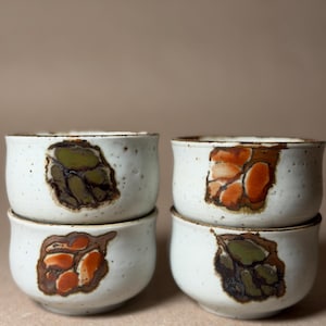 May include: Four white ceramic bowls with a speckled texture. Each bowl features a unique abstract design in shades of brown, orange, and green. The bowls are stacked in pairs, showcasing their handmade quality and earthy aesthetic.