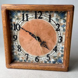 May include: A square wooden clock with a central peach-colored circular face featuring a swirling texture. The clock has black numerals from 1 to 12 and ornate black hands. A decorative border of blue and white floral patterns surrounds the center.
