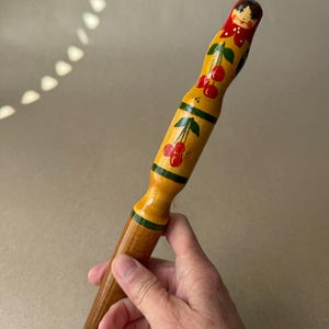 May include: A wooden rolling pin with a painted design. The handle is painted yellow with red cherries and green leaves. The top of the rolling pin has a painted face. The bottom is a natural wood color.