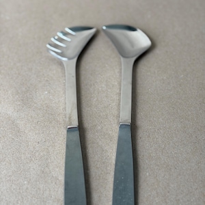 May include: Two stainless steel salad servers. One server has a slotted head, the other a solid, curved head. Both have long, flat handles. The servers are displayed against a neutral background.