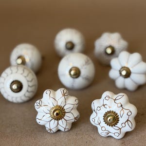 May include: A collection of eight ceramic cabinet knobs in white with various gold-toned patterns and metallic centers. The knobs feature round and scalloped shapes with intricate line work, including floral motifs and geometric designs.