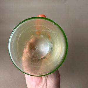 May include: A clear glass with a green rim and an orange stopper. The glass has a slightly yellow tint and appears to be empty. The image is taken from a top-down perspective, showcasing the inside of the glass.