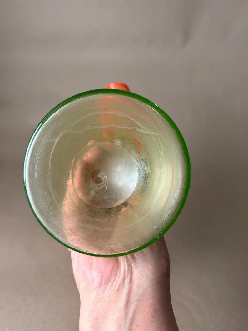 May include: A clear glass with a green rim and an orange stopper. The glass has a slightly yellow tint and appears to be empty. The image is taken from a top-down perspective, showcasing the inside of the glass.