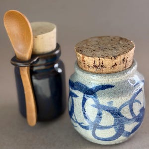 May include: A small, light gray ceramic jar with blue circular patterns and a cork lid, positioned next to a dark blue ceramic jar featuring a side loop that holds a small wooden spoon with a light brown finish.