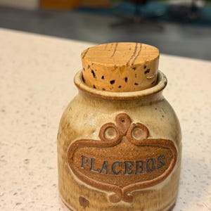 May include: A small, tan ceramic jar with a cork stopper. The jar has a brown, decorative label that reads "PLACEBOS". The jar has a rustic, handmade appearance.