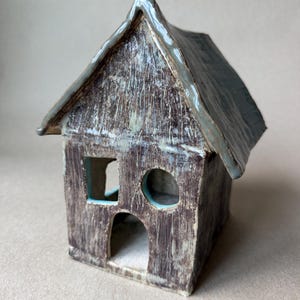 May include: A handcrafted ceramic house with a textured, weathered appearance. The miniature house features a dark brown exterior with blue accents around the windows and door. The roof is a darker shade of blue. The house has a rustic, artisanal aesthetic.