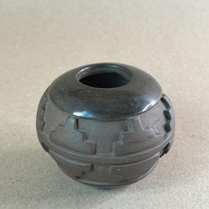 May include: A small, round, dark brown ceramic pot with a smooth, polished rim and a matte, textured body featuring carved geometric step patterns encircling the exterior. The vessel has a wide, circular opening at the top and a rounded base.