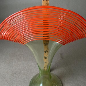 May include: A clear glass vase with a flared top and a round base. The top is adorned with bright orange, curved lines. A wooden ruler is placed inside the vase, measuring approximately 10 inches in height. The vase has a green tint.