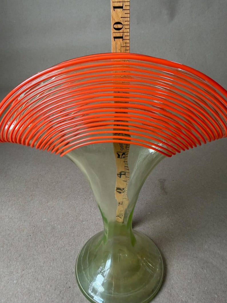 May include: A clear glass vase with a flared top and a round base. The top is adorned with bright orange, curved lines. A wooden ruler is placed inside the vase, measuring approximately 10 inches in height. The vase has a green tint.