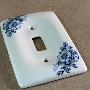 May include: A rectangular white light switch wall plate featuring a blue floral pattern printed on the top left and bottom right corners. The plate has a central rectangular cutout for a toggle switch and two circular screw holes for mounting.