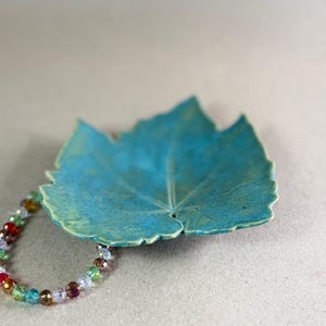 May include: A teal ceramic leaf-shaped dish with visible veining and a textured surface, accompanied by a circular bracelet made of small, multi-colored, translucent glass beads in shades of red, green, blue, and yellow.