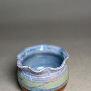 May include: A handmade ceramic bowl with a wavy rim. The bowl features a blend of colors, including blue, green, and brown. The exterior has a textured appearance, and the interior is a solid blue. This decorative bowl is suitable for serving or display.