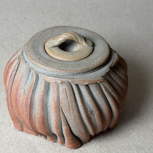 May include: A handcrafted ceramic jar with a lid. The jar has a textured, pleated design in shades of brown and gray. The lid features a loop handle. This decorative piece is suitable for home decor.