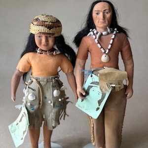 May include: Two Native American dolls. One doll wears an orange top, a skirt adorned with shells, and a woven hat. The other doll wears an orange top and brown pants, with a shell necklace. Both dolls hold a green paper.