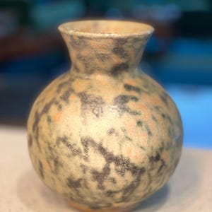 May include: A small, round ceramic vase with a speckled pattern. The vase has a cream-colored base with dark gray and peach-colored spots. The vase has a narrow neck and a wide, rounded body. The overall aesthetic is rustic and handmade.