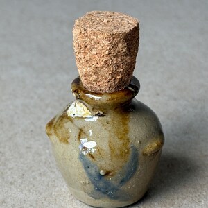 May include: A small, rounded ceramic bottle with a glossy, mottled tan and brown glaze featuring a dark blue accent mark. The bottle has a short, narrow neck and is sealed with a cylindrical, textured natural cork stopper at the top.