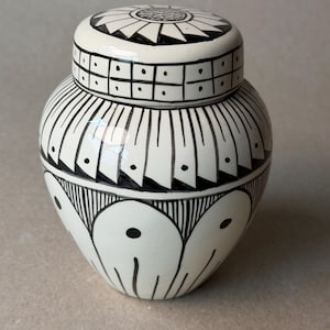 May include: A white ceramic jar with a lid, adorned with black geometric patterns. The jar features a rounded body and a lid with a circular design. The black patterns include lines, dots, and square shapes, creating a striking contrast.