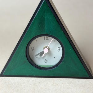 May include: A triangular desk clock with a dark green wooden frame and a circular face. The clock face is light gray with black numerals at 3, 6, 9, and 12 o'clock. It features black hour and minute hands and a thin pink second hand.