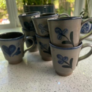 May include: A collection of beige ceramic mugs with blue floral designs and handles. The mugs have a dark blue rim and a heart design on one mug. The mugs are stacked and arranged on a light-colored surface.