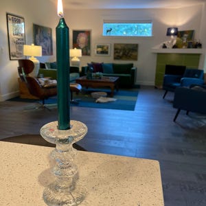 May include: A lit, emerald green candle in a clear, textured glass candlestick. The candle is tall and slender, with a bright flame. The candlestick has a detailed, layered design. The background shows a living room with art and furniture.