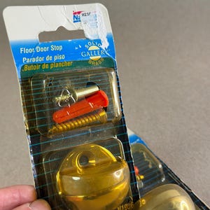 May include: A floor door stop in retail packaging labeled Solid Gallery Brass. The package includes a gold-colored metal door stop, a gold-colored screw, and an orange plastic wall anchor. The text on the packaging reads Floor Door Stop.