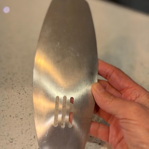 May include: A stainless steel spatula with a rectangular shape and rounded top. The spatula has three vertical slots near the bottom. The spatula is held by a person's hand.