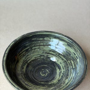 May include: A handmade ceramic bowl with a dark green and black spiral design. The bowl has a glossy finish and a slightly rough texture around the rim. The interior features a dark, swirling pattern.