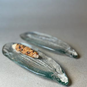 May include: Two elongated, clear glass dishes shaped like corn husks with textured, rippled surfaces. One dish contains a small, realistic miniature cob of corn with yellow and brown kernels, while the other dish is empty and displays the same glass form.