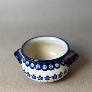 May include: A ceramic bowl with two handles, featuring a cream-colored base decorated with a blue and green peacock-eye pattern around the rim and a repeating blue floral design on the lower body, finished with a dark blue rim and handle accents.