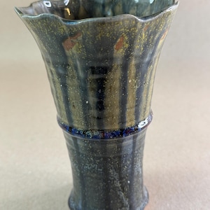 May include: A ceramic vase with a flared, wavy rim and a tapered body. The surface features a dark, mottled glaze in shades of charcoal, olive green, and deep blue, with vertical streaking and subtle orange-brown speckles throughout the exterior.