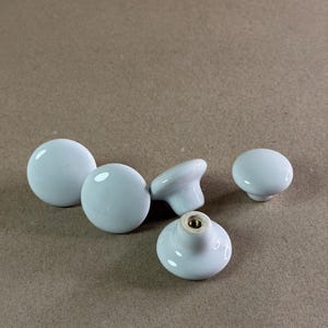 May include: A set of four smooth, glossy white ceramic cabinet knobs with a rounded, mushroom-shaped profile. Each knob features a central brass-colored metal insert at the base for mounting, showcasing a clean, reflective surface finish.
